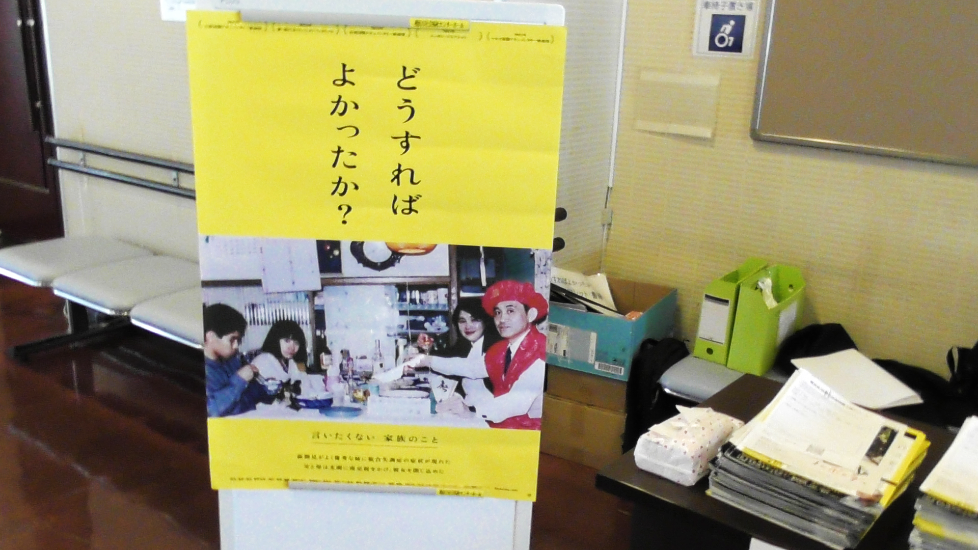 通路には作品のポスターがあり、黄色背景に黒文字で「どうすればよかったか？」と書かれ、下部には当事者たちの写真が掲載されている。