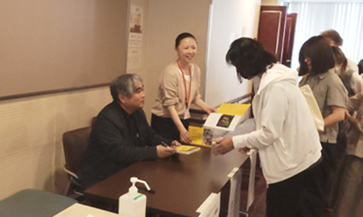 販売コーナーで著書やパンフレットを購入した来場者に、監督が著書へサインを書いている様子。